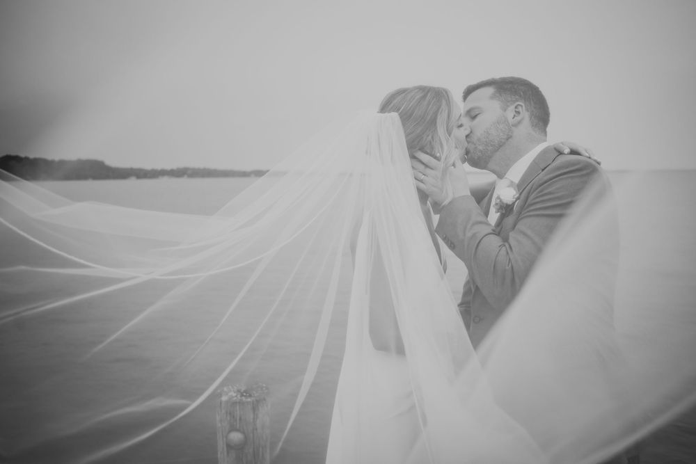 Black and white artistic wedding photography showing couple kissing under flowing bridal veil against scenic backdrop.
