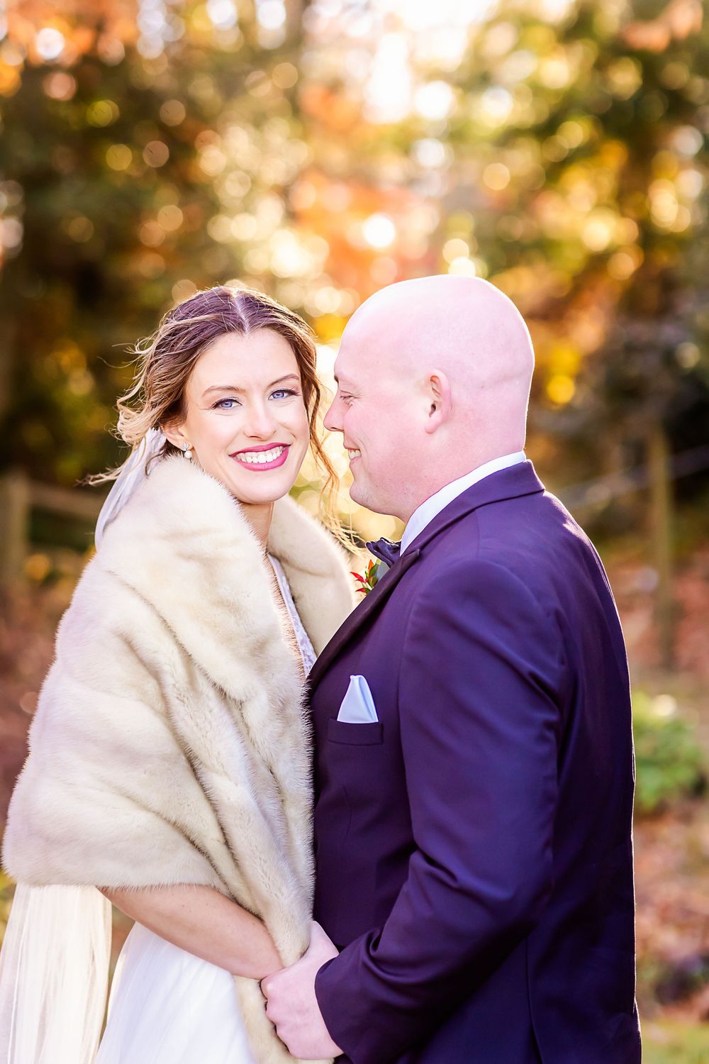 A couple in wedding attire share an intimate moment outdoors, with the bride wearing a cream fur wrap and elegant dress.
