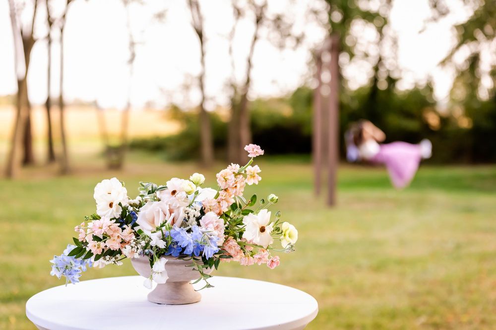 Floral centerpiece arrangement in pastel colors sits on white round table outdoors with blurred garden background.