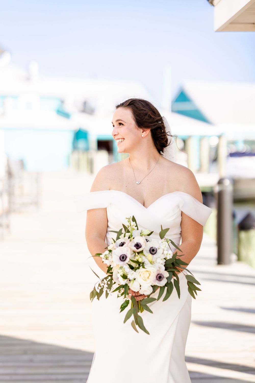 Bride in white off-shoulder wedding dress smiles holding white and blush toned anemone bouquet with greenery outdoors.