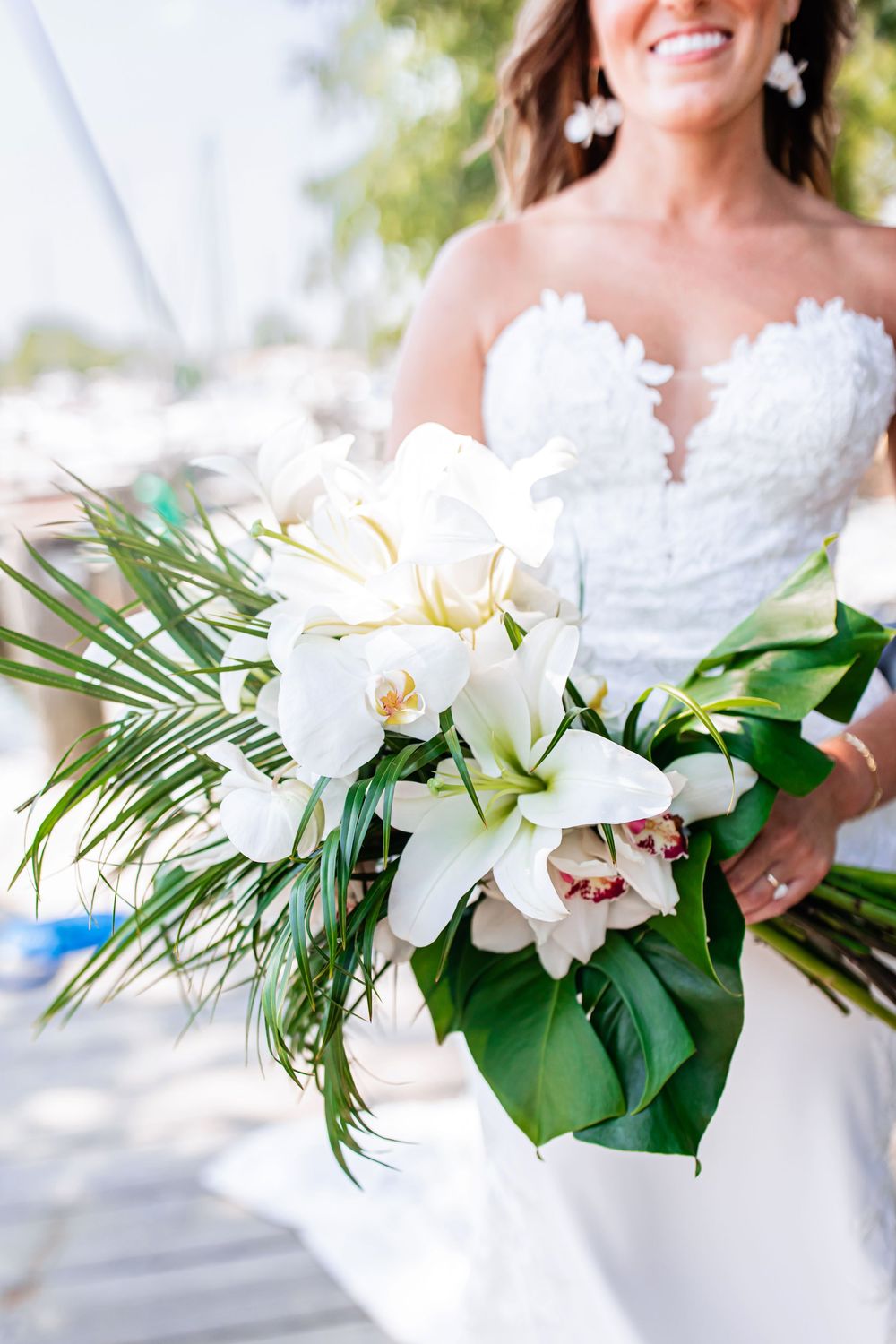Elegant white bridal bouquet features orchids and palm fronds against a white wedding dress in natural light.