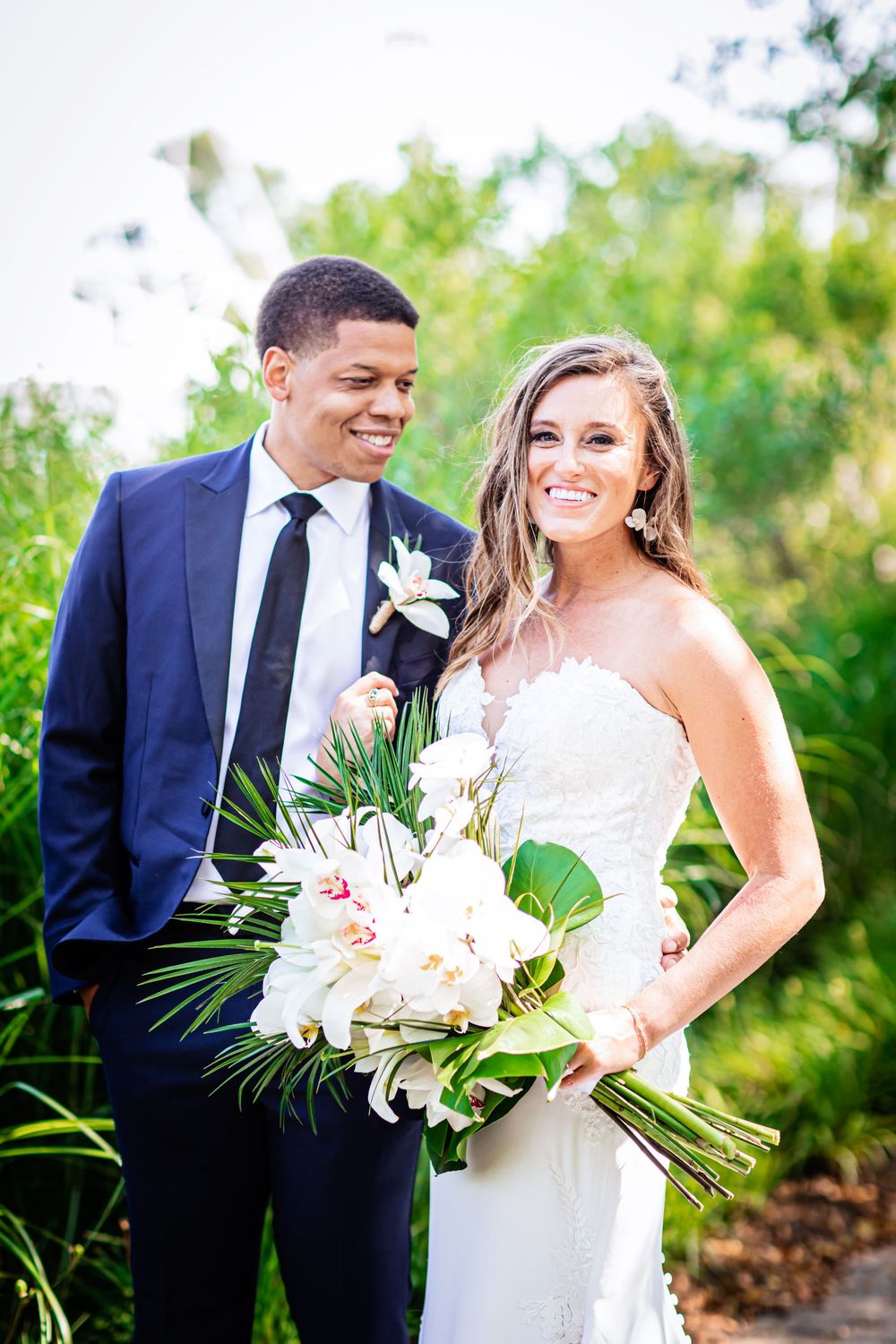 A happy couple on their wedding day poses outdoors with a tropical bouquet of white flowers and greenery.