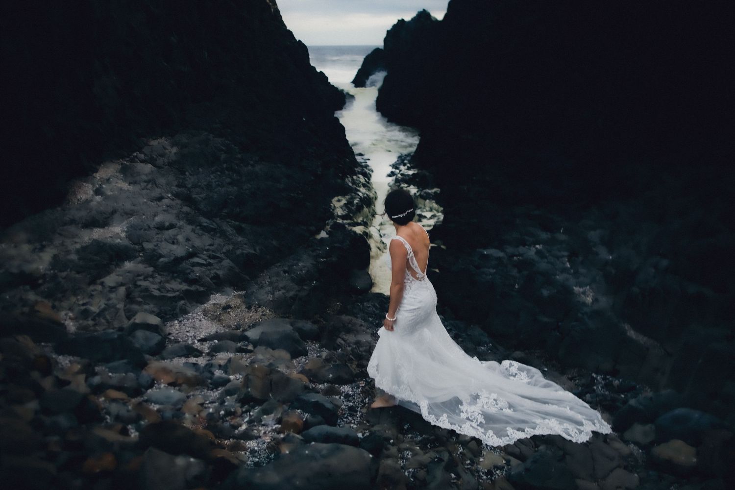 A flowing white wedding dress contrasts dramatically against dark rocky cliffs at a moody coastal location.
