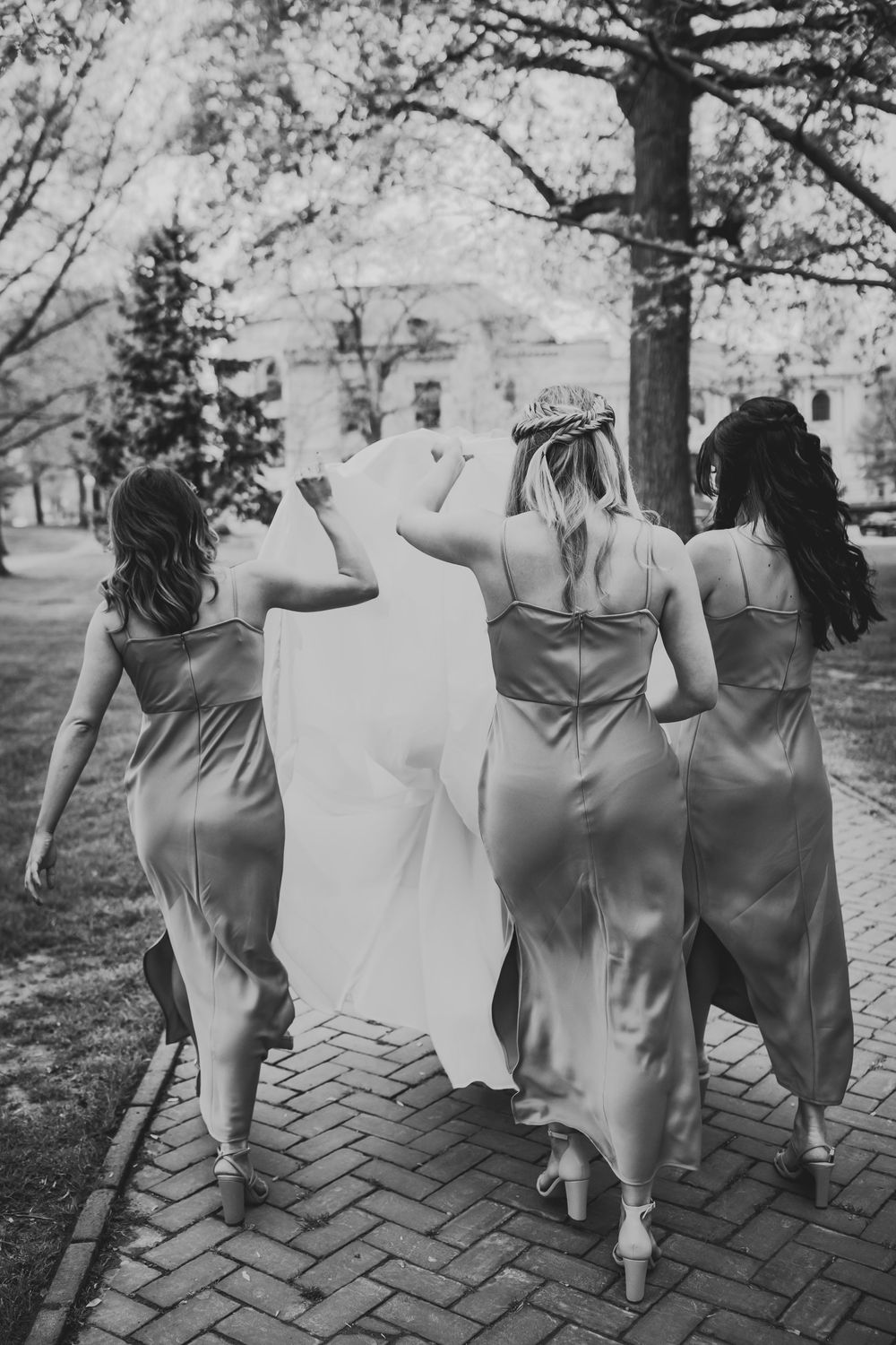 Black and white photo of bridesmaids walking down brick path in elegant dresses, taken from behind.