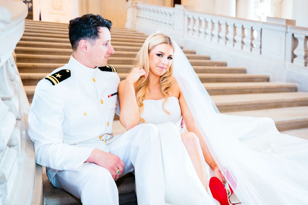 A bride in a white gown and military groom in dress uniform sit together on elegant marble steps.
