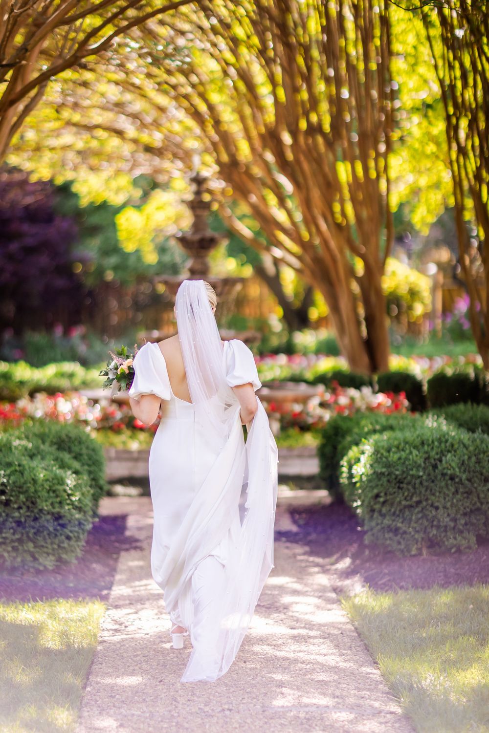 A person in a flowing white wedding dress walks down a garden path beneath arching trees on a sunny afternoon.