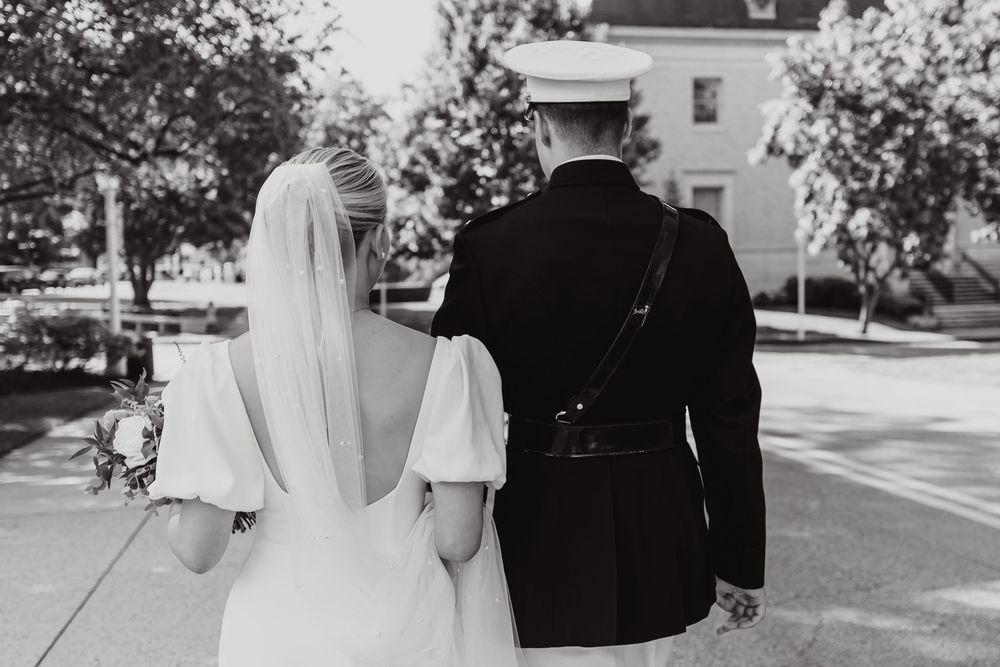 Black and white photograph capturing the rear view of a bride in a white dress and groom in military uniform walking together.