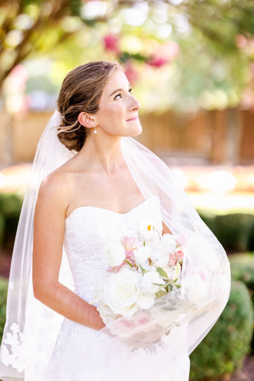 A bride in white wedding dress looks upward while holding a bouquet in an outdoor garden setting.