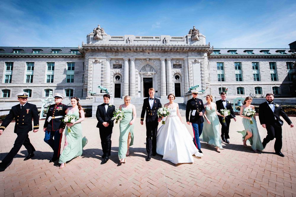 A wedding party in formal attire walks hand in hand along a pathway outside an elegant white historic building.