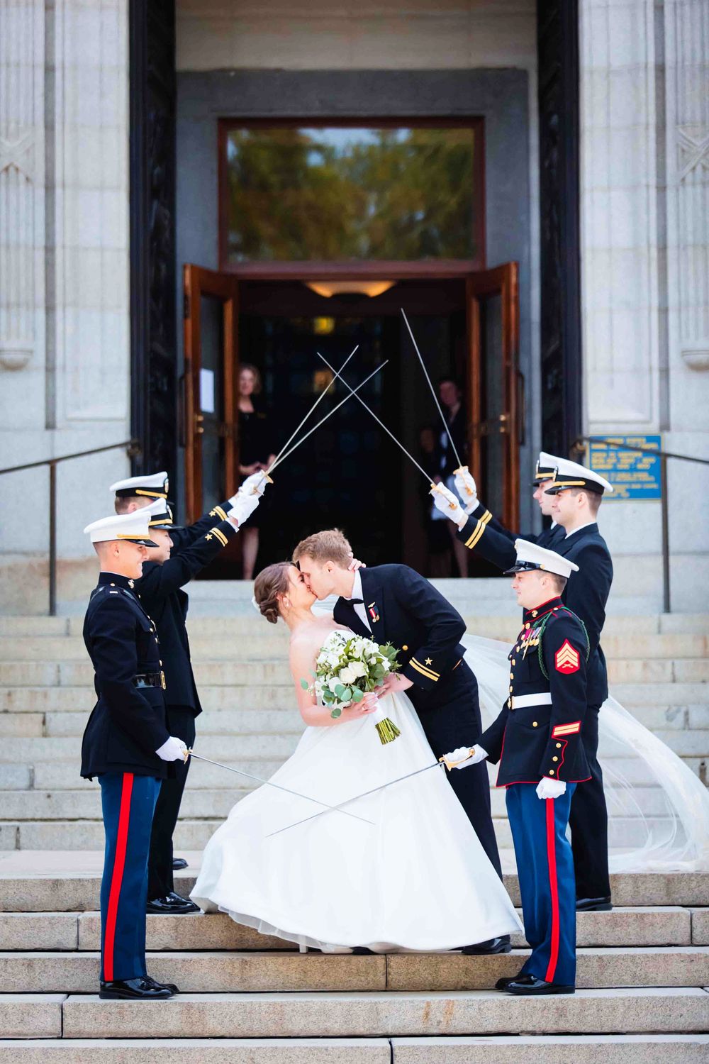 Military service members in dress uniforms create sword arch as newlywed couple shares romantic kiss on church steps.