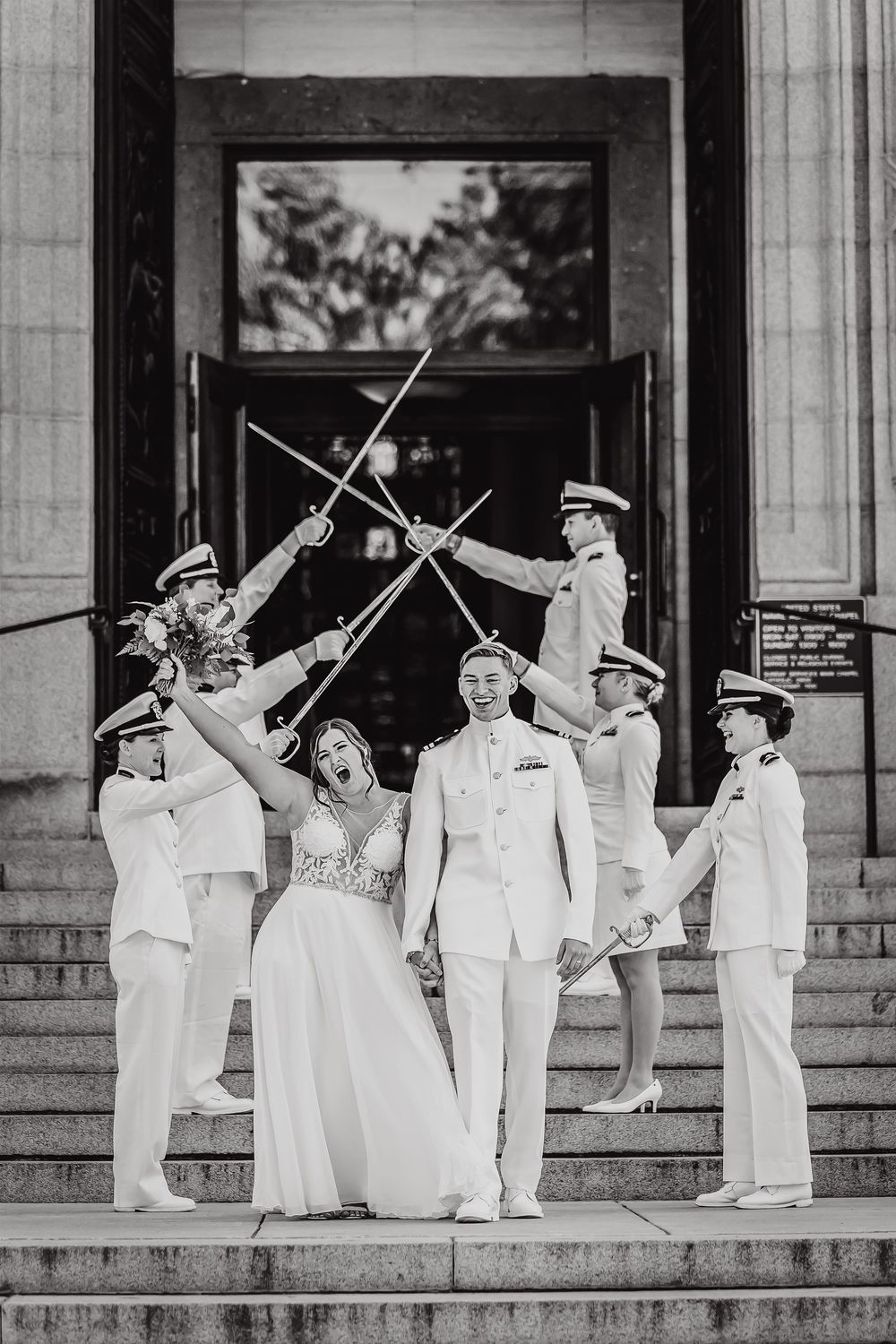 A bride and groom stand on stairs with military officers forming a traditional sword arch in black and white photography.