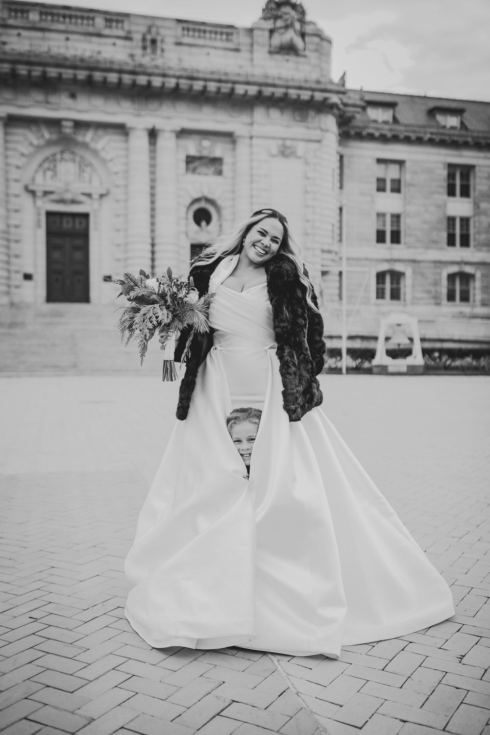 A bride in a flowing white gown holds a bouquet while standing outside a historic building in black and white photography.