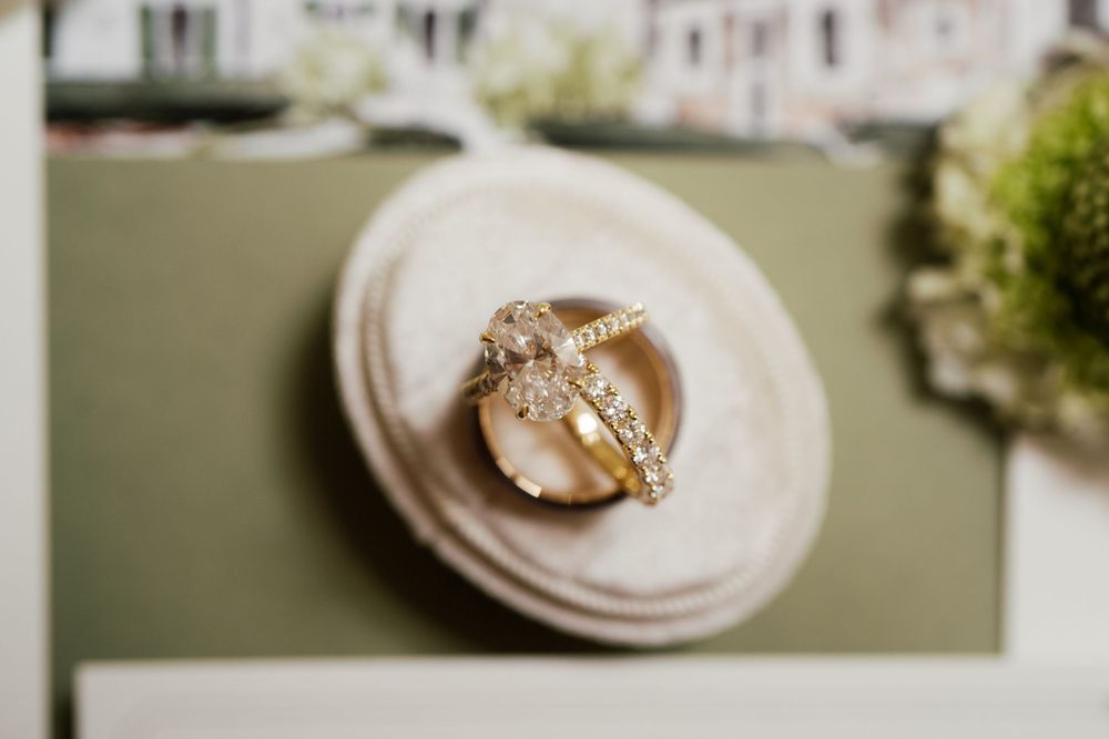 A sparkling diamond engagement ring rests on a cream-colored textured surface with soft background bokeh.