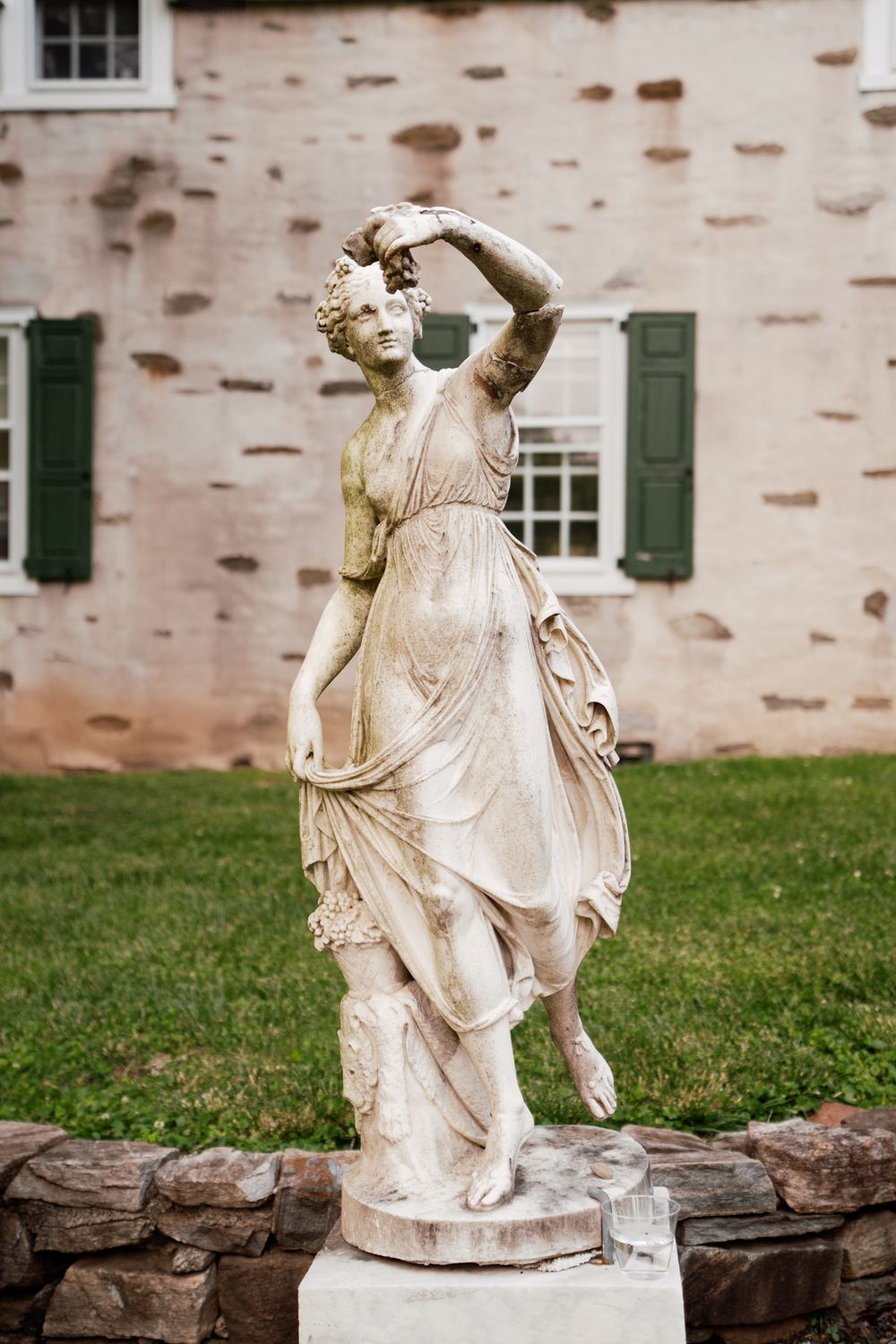 Classical white stone garden statue of a dancing female figure set against pink stone building with green shutters.