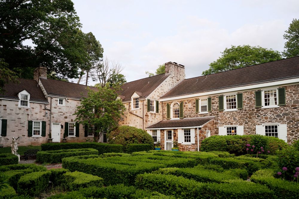 Historic stone colonial house with manicured hedge maze garden and traditional shuttered windows in countryside setting.
