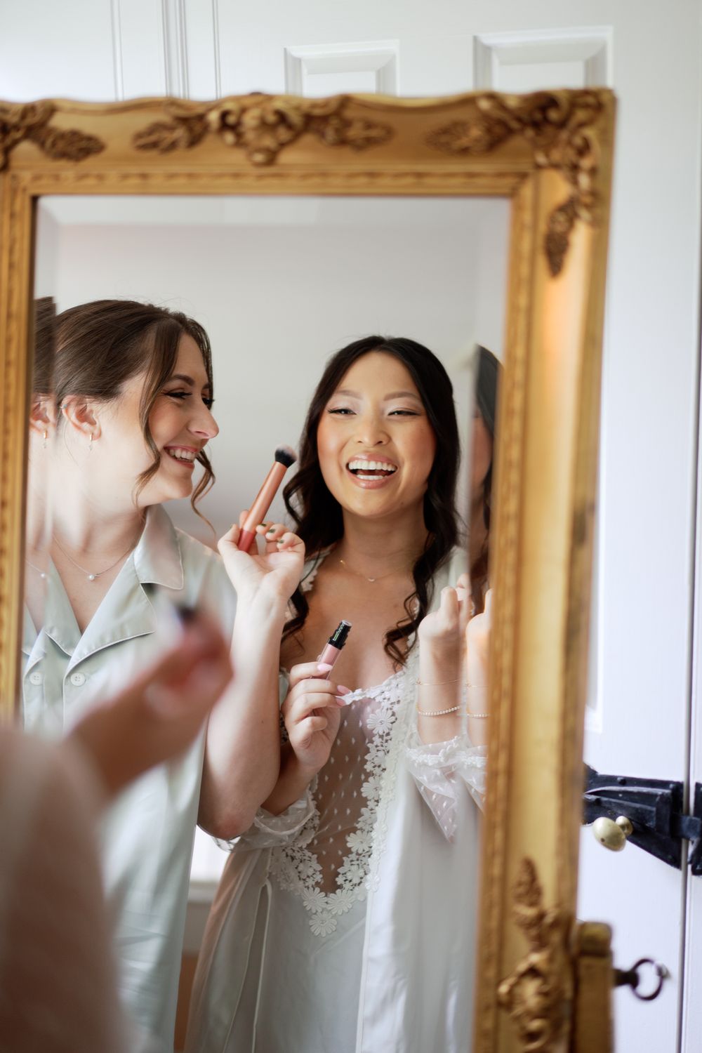 Two friends share a joyful moment applying makeup together while looking in an ornate gold-framed mirror.