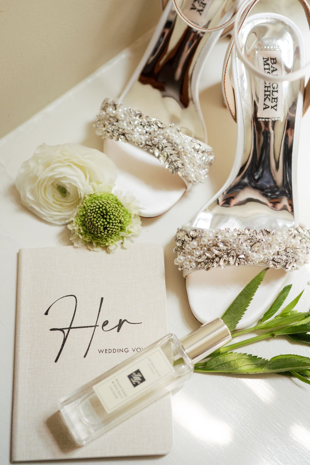 Elegant wedding details including white heels with crystal embellishments, a Jo Malone perfume bottle, and delicate floral accents.