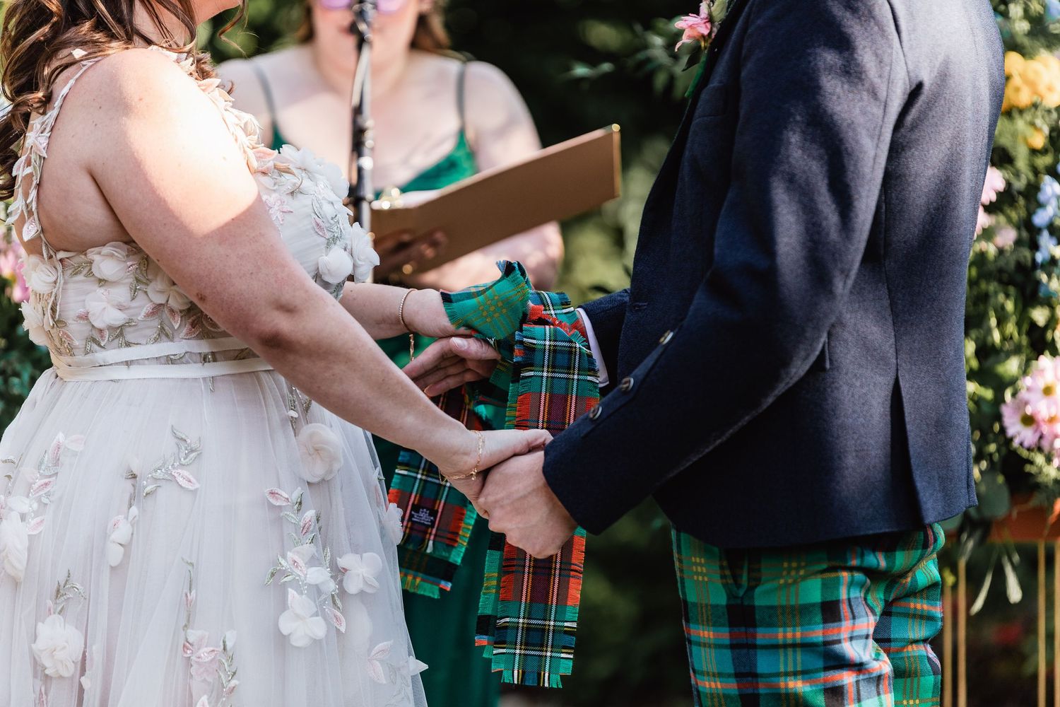 Two people hold hands during a wedding ceremony, with the groom wearing a traditional green and blue tartan kilt.