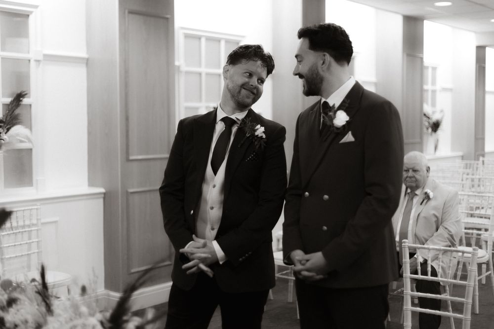 Two grooms share a lighthearted moment together during their wedding ceremony while wearing formal black suits.