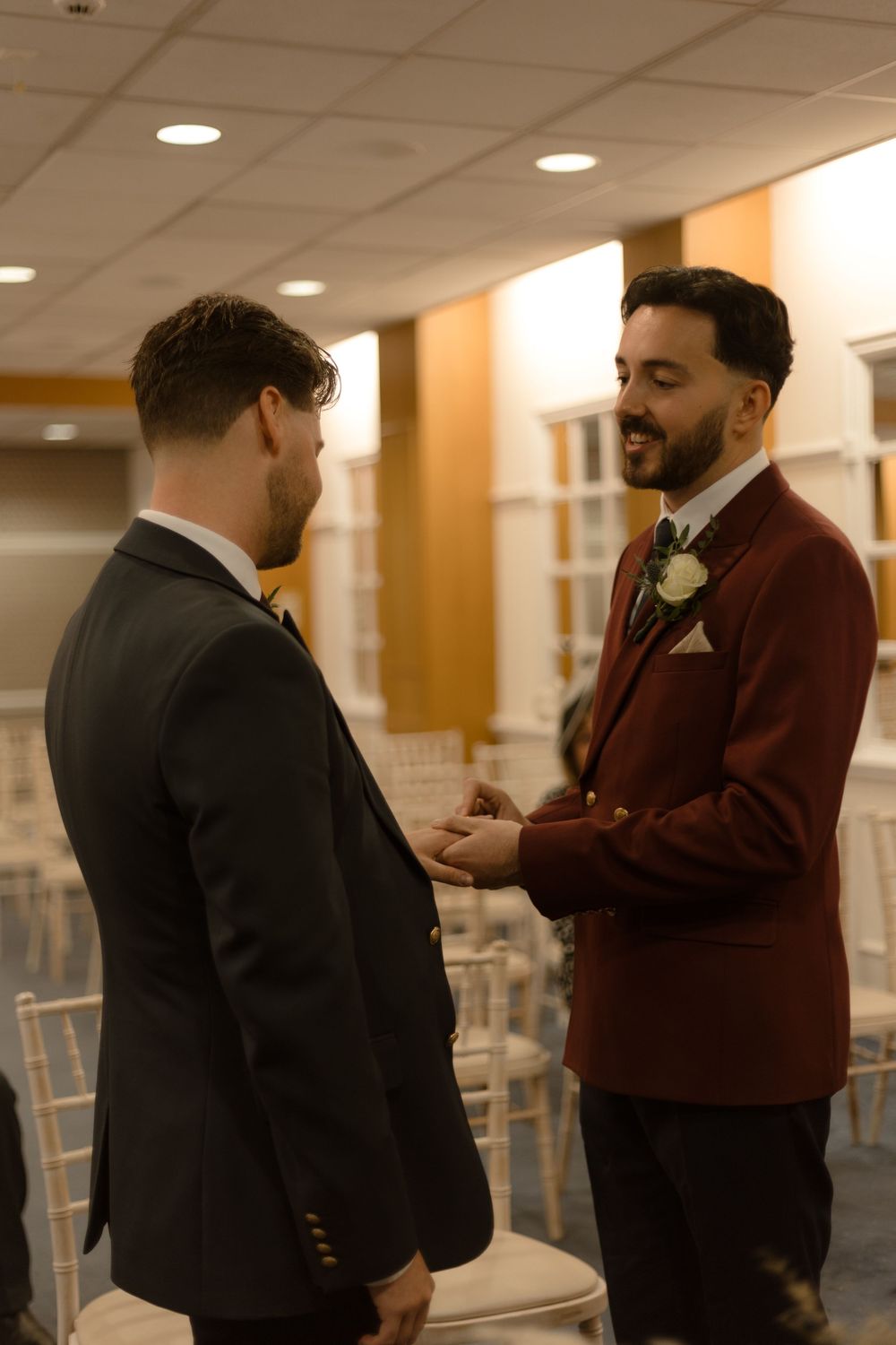 Two people in formal attire having a conversation in a hallway with recessed lighting and chairs in the background.