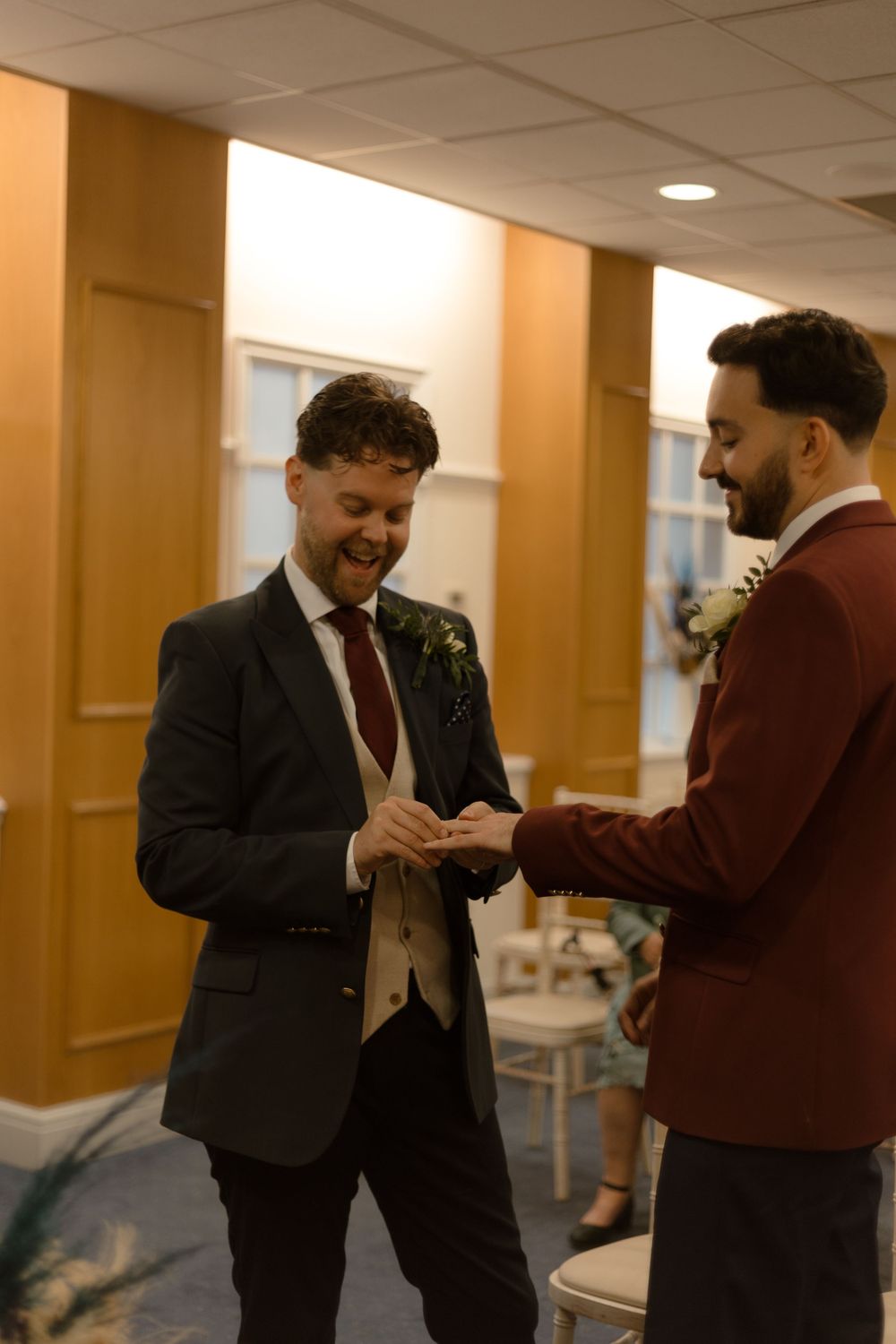 Two people in formal attire conversing and laughing together in an indoor hallway with warm lighting.