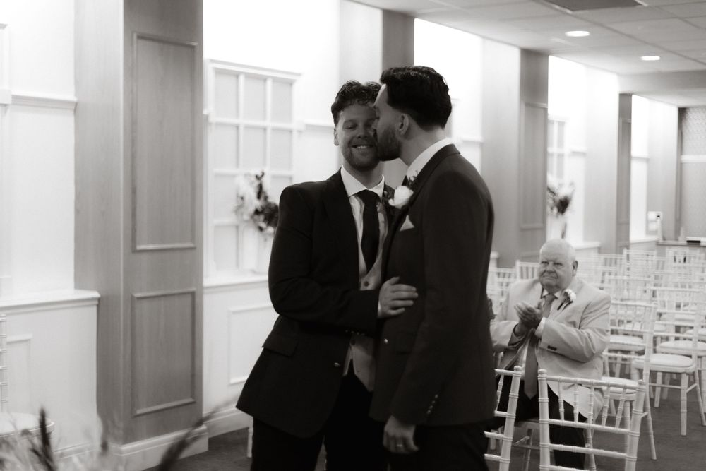 Two people in black suits share an intimate moment during a wedding celebration in a bright indoor venue with white chairs.