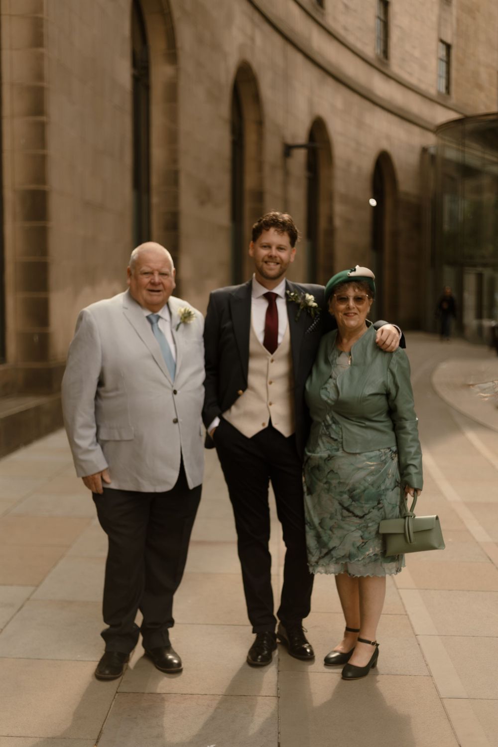 Three people in formal attire pose together on a stone-paved walkway outside a grand building with arched windows.