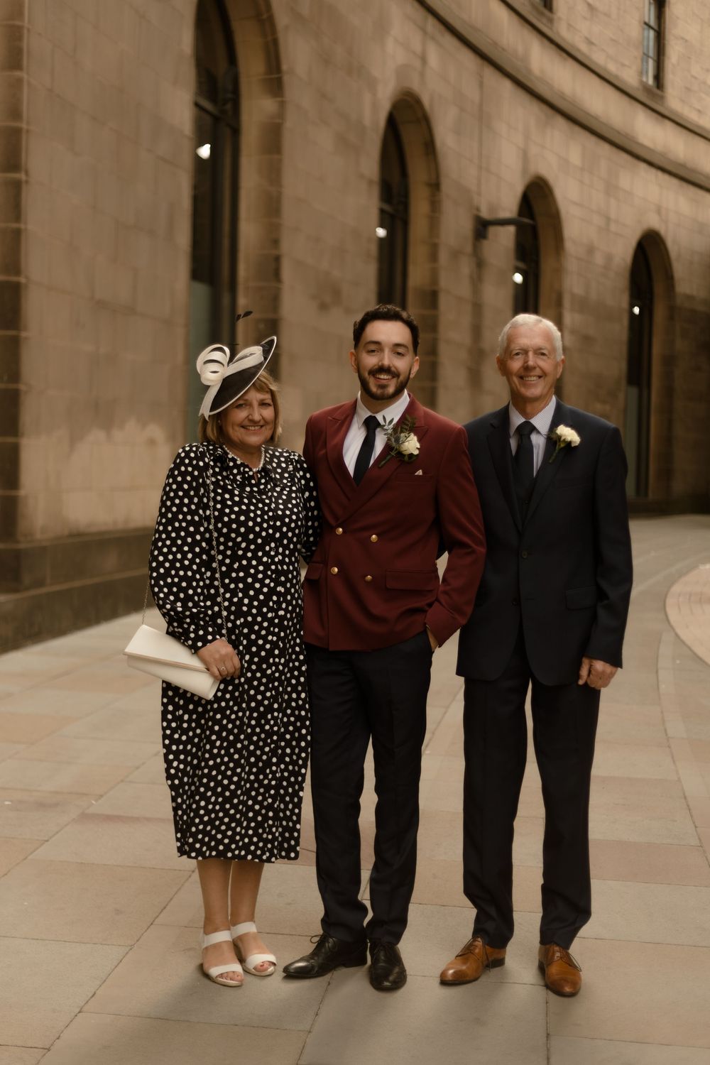 A group of people stands in a row together wearing formal attire along a stone building wall.