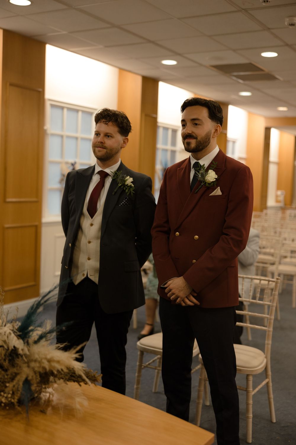 Two grooms stand together at a wedding venue wearing coordinating suits, one in burgundy and one in black with a light vest.
