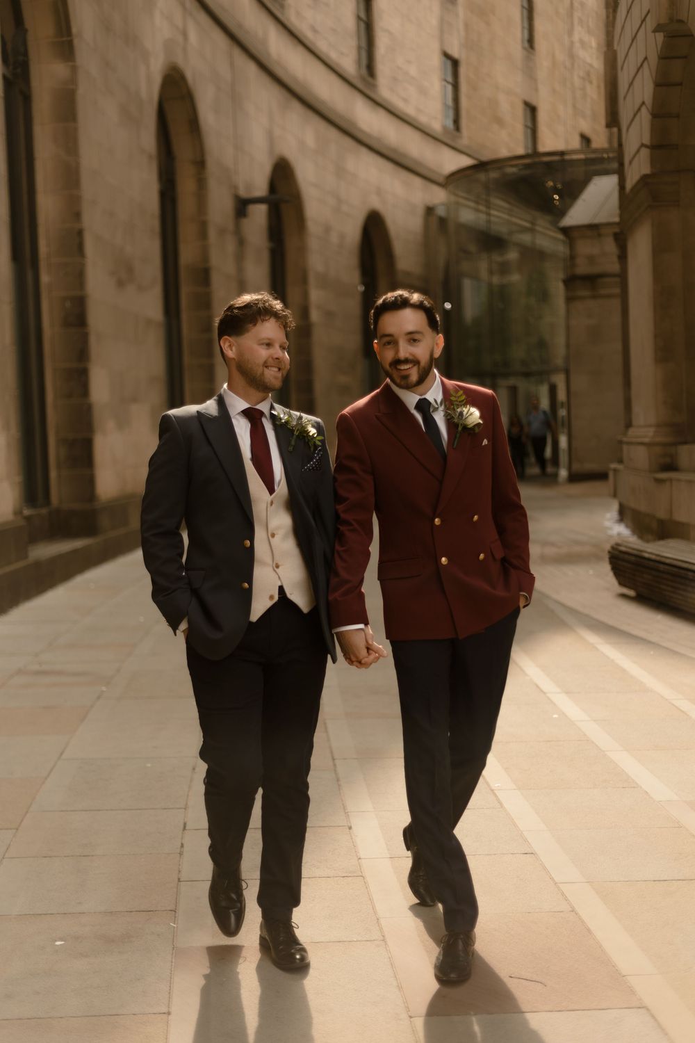 Two grooms walking hand in hand down a grand hallway, wearing formal attire in burgundy and black with boutonnières.