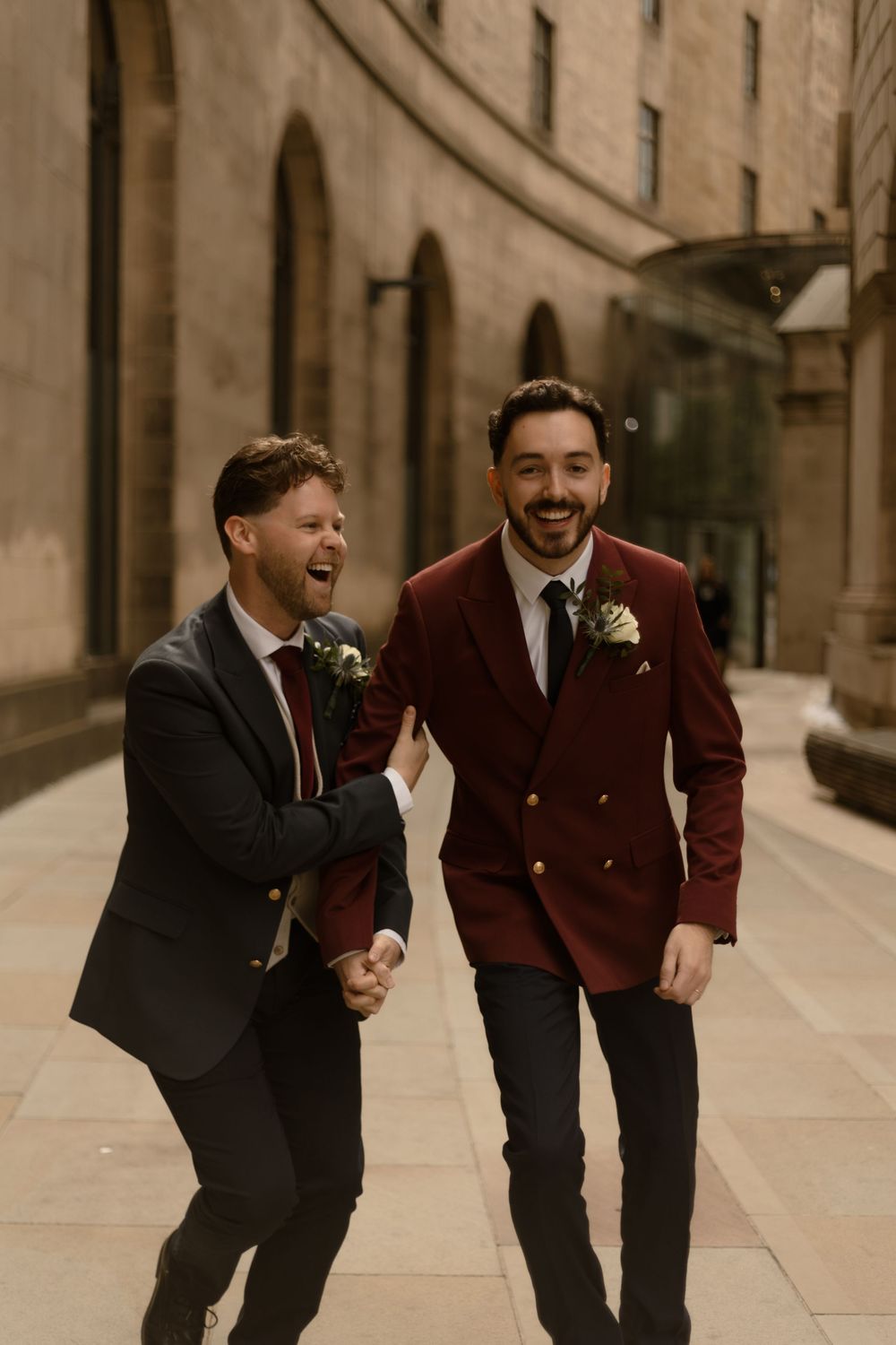 Two people in formal suits with boutonnieres share a candid laughing moment while walking along a stone building.