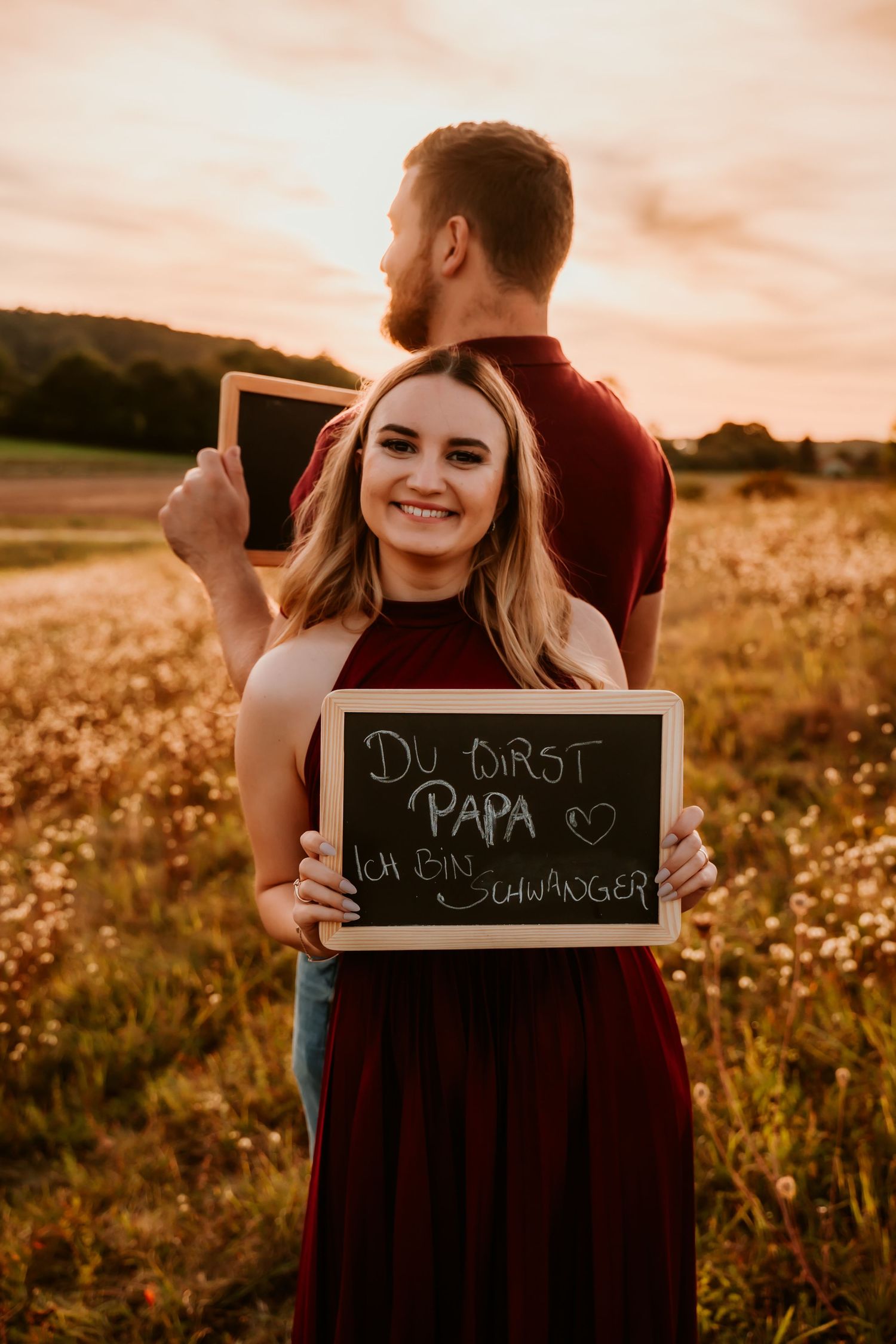 Two people hold chalkboards in a golden field during sunset while wearing burgundy clothing.
