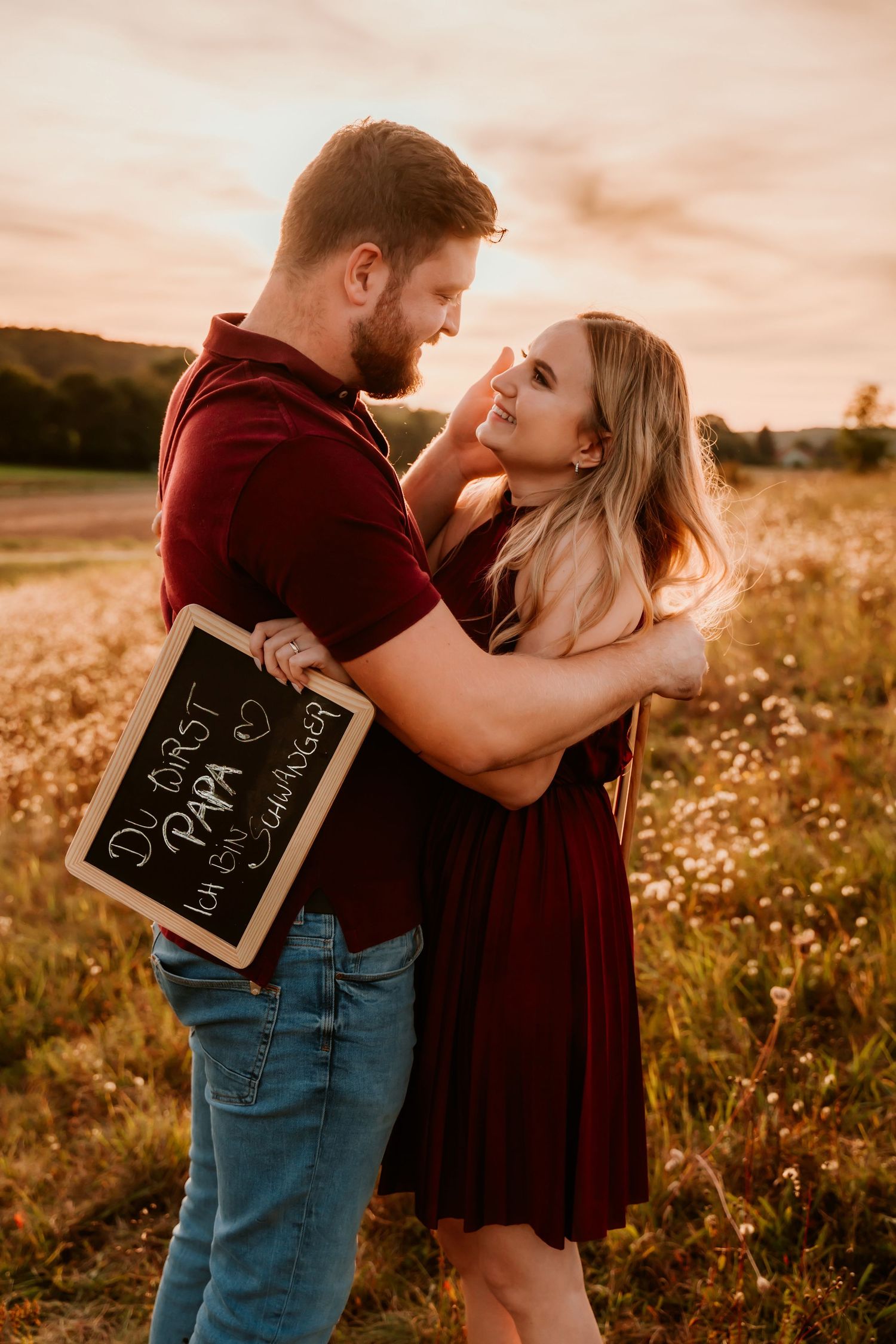 A romantic couple embrace while holding a chalkboard sign in a sunset-lit meadow.