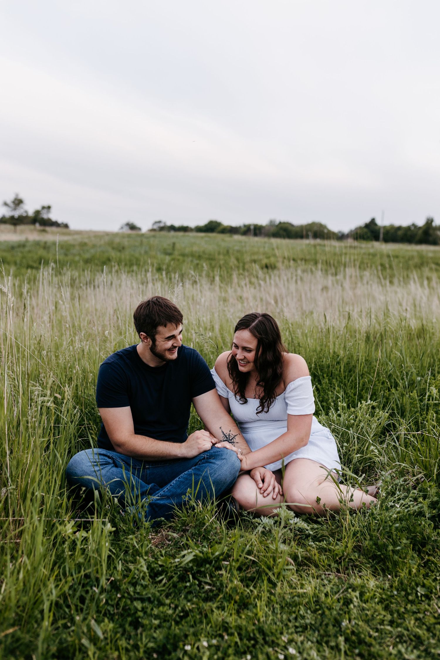 A couple sits together in a grassy field on an overcast summer day, smiling and looking at each other.