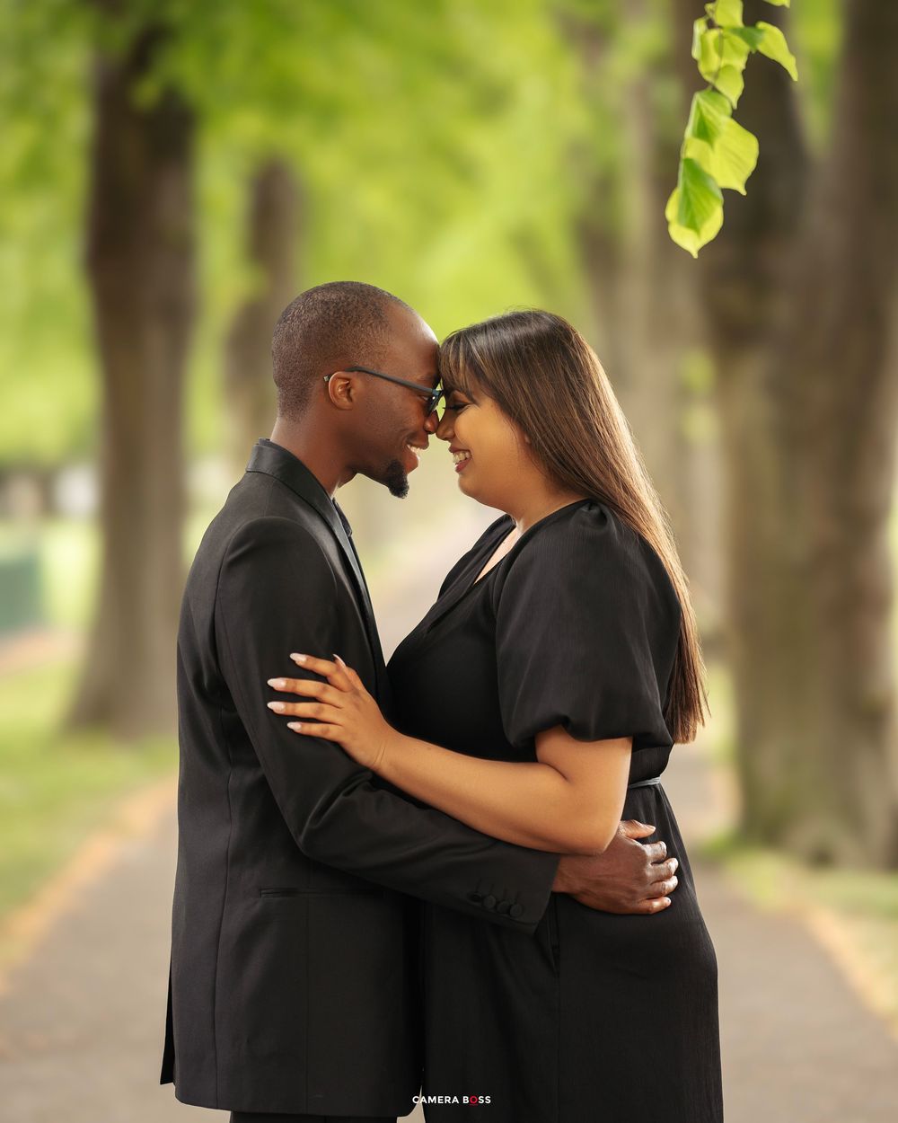 A romantic couple shares an intimate moment on a tree-lined path in the park, both wearing elegant black outfits.