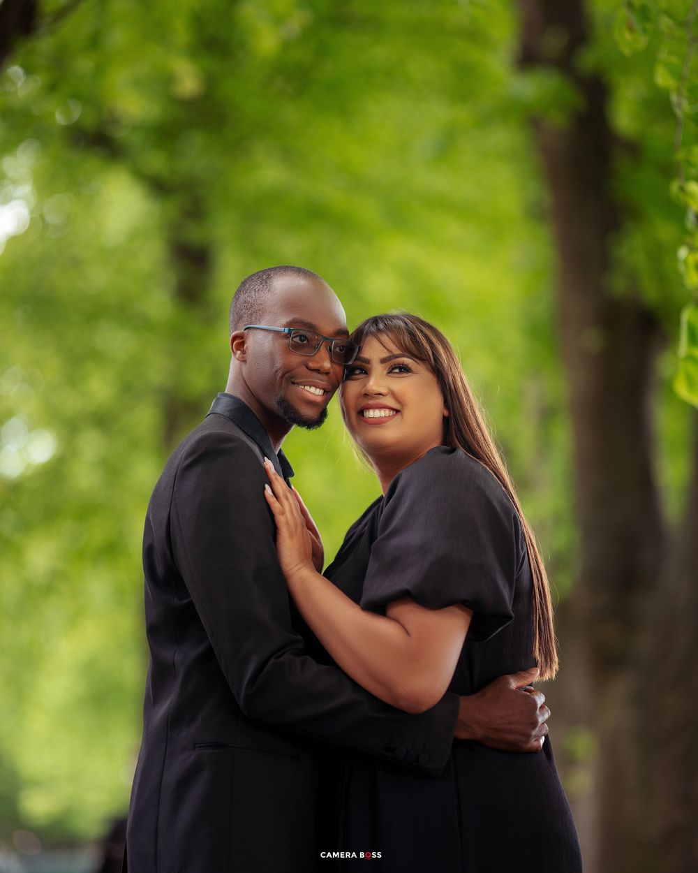 A loving couple embraces outdoors while wearing matching black outfits against a green natural background.