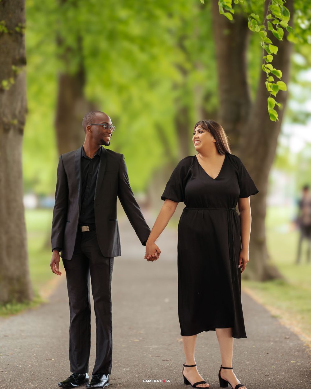 A couple wearing black formal attire holds hands while walking along a tree-lined path on a sunny day.