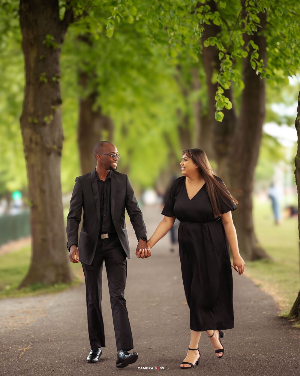 A happy couple walks hand in hand down a picturesque tree-lined pathway while wearing matching black formal attire.