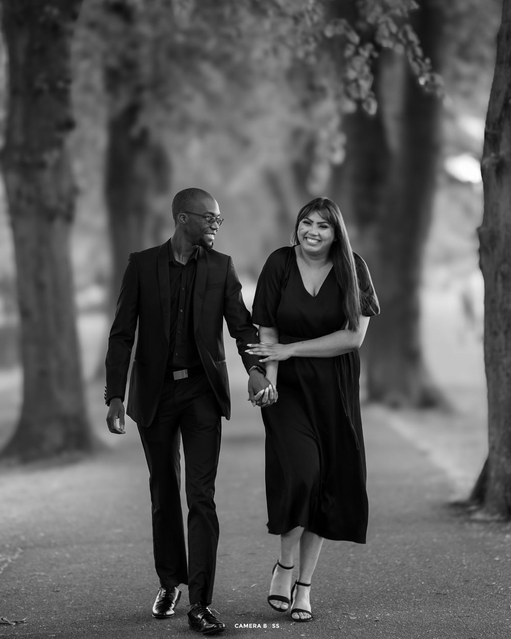 Couple in black attire walking together and laughing on a tree-lined path in black and white photography.