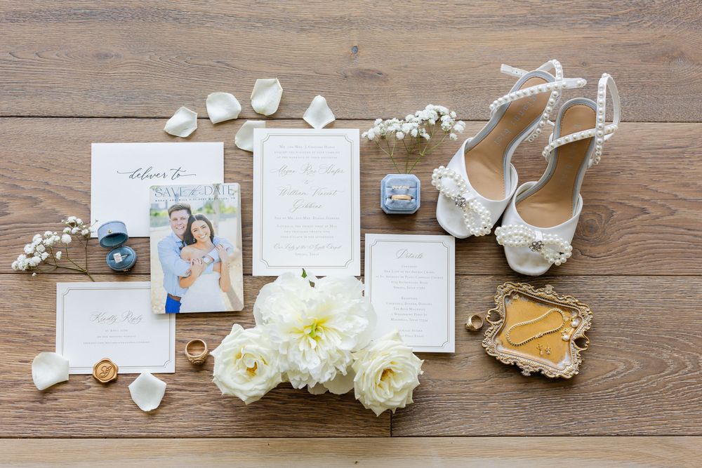 Wedding details flatlay with invitations, white flowers, and bridal shoes on wooden background.