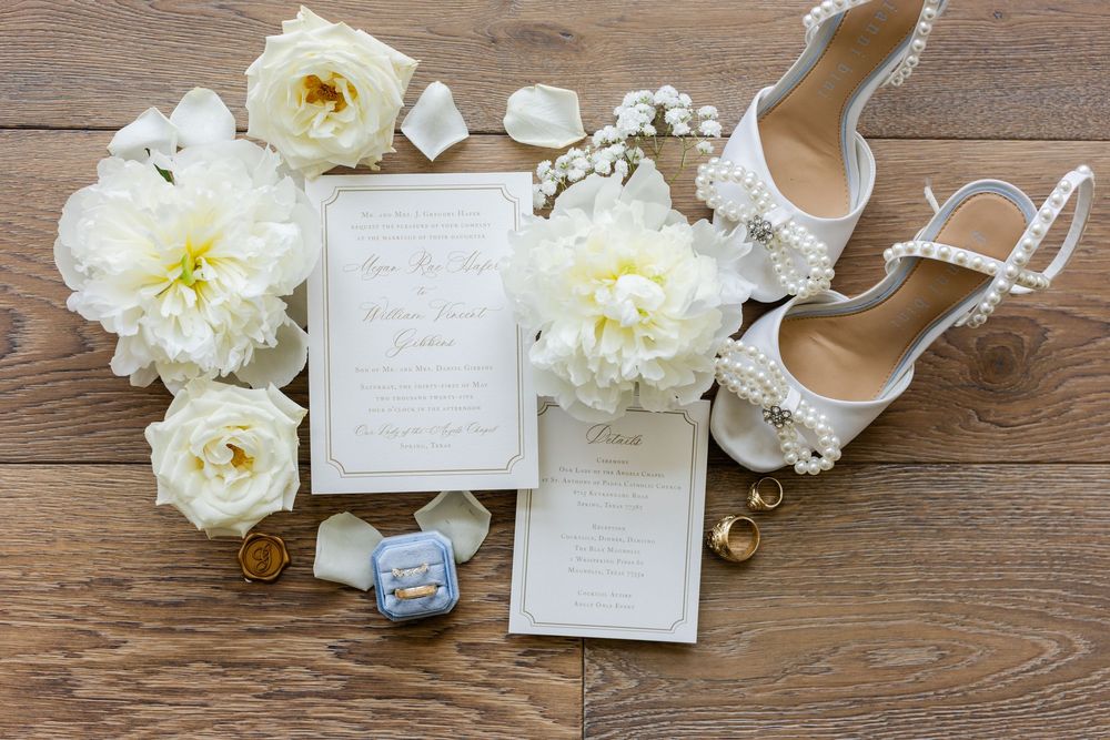 Wedding stationery with white peonies and bridal shoes arranged on wooden surface.