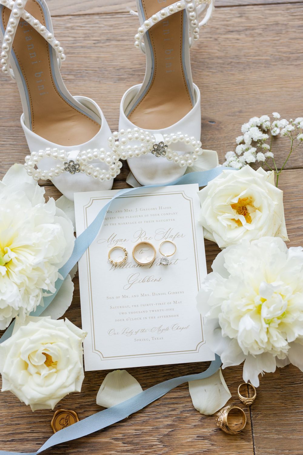 Wedding details flat lay with white heels, rings, invitation and white roses on wooden surface.