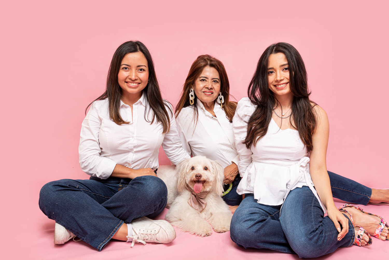 Three people in white tops and blue jeans sit together with a small white dog on a pink backdrop.