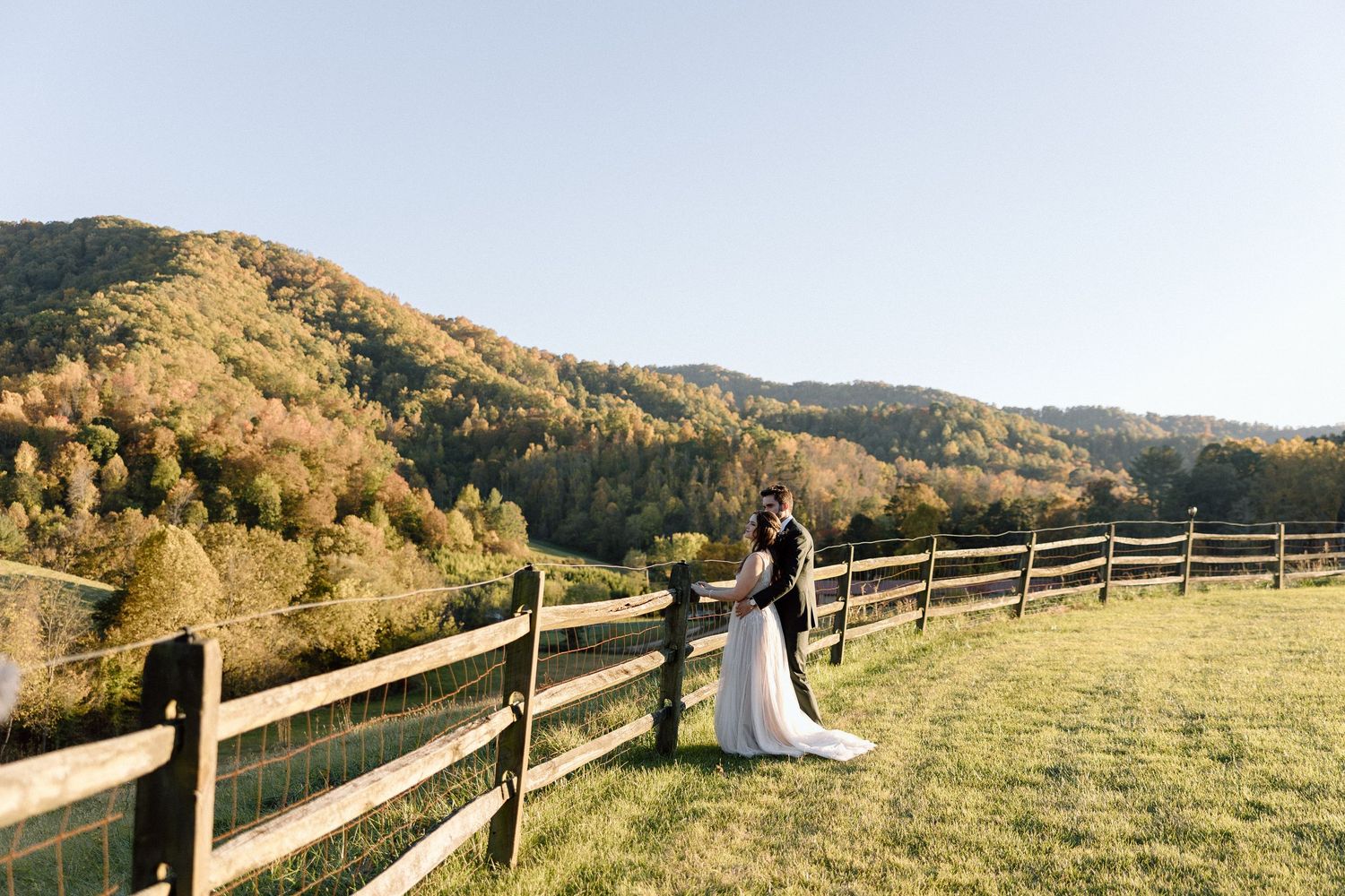 Two figures in wedding attire embrace near wooden fence overlooking mountain landscape during golden hour at countryside.