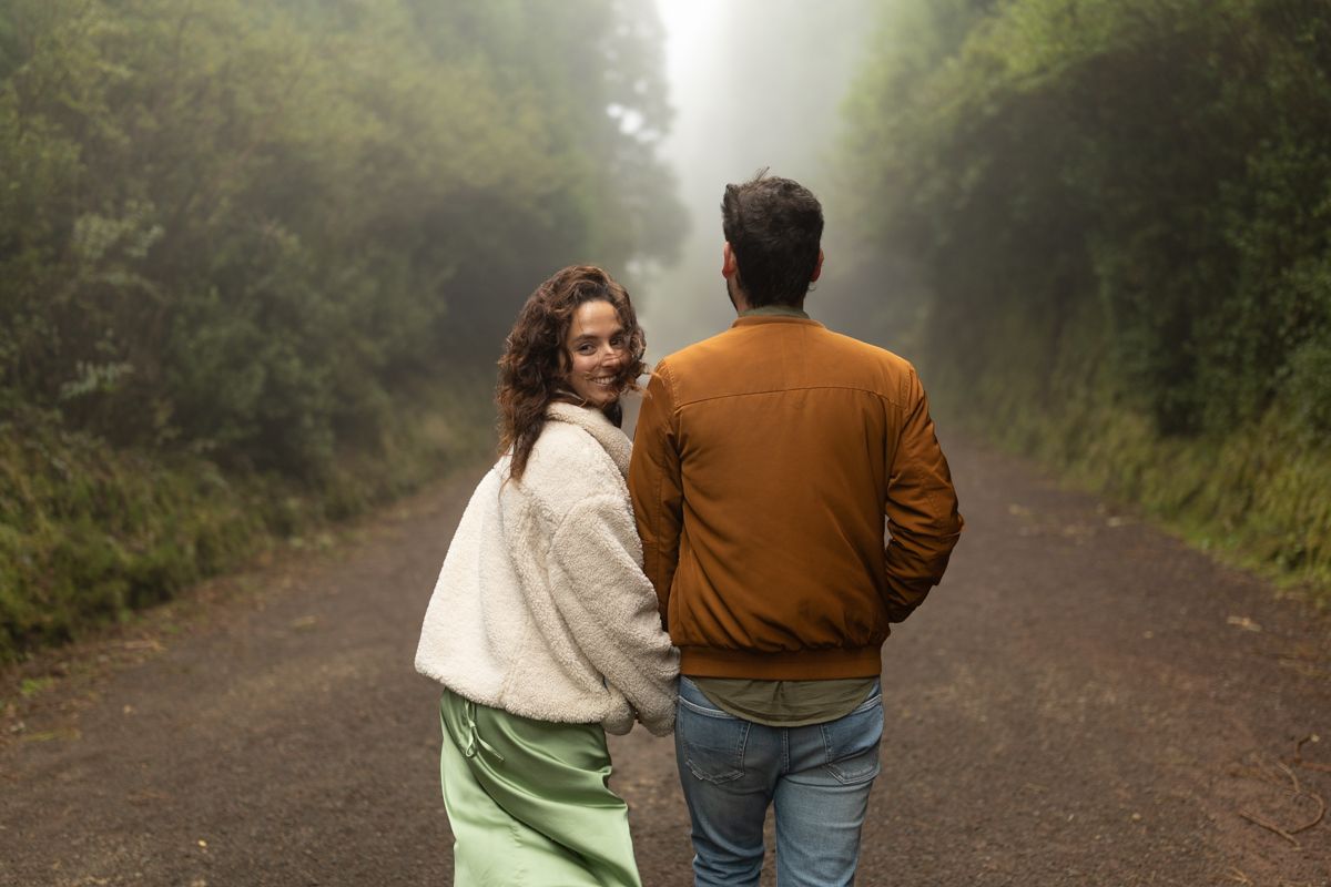 A couple walking down a misty forest road as the woman looks back over her shoulder at the camera.