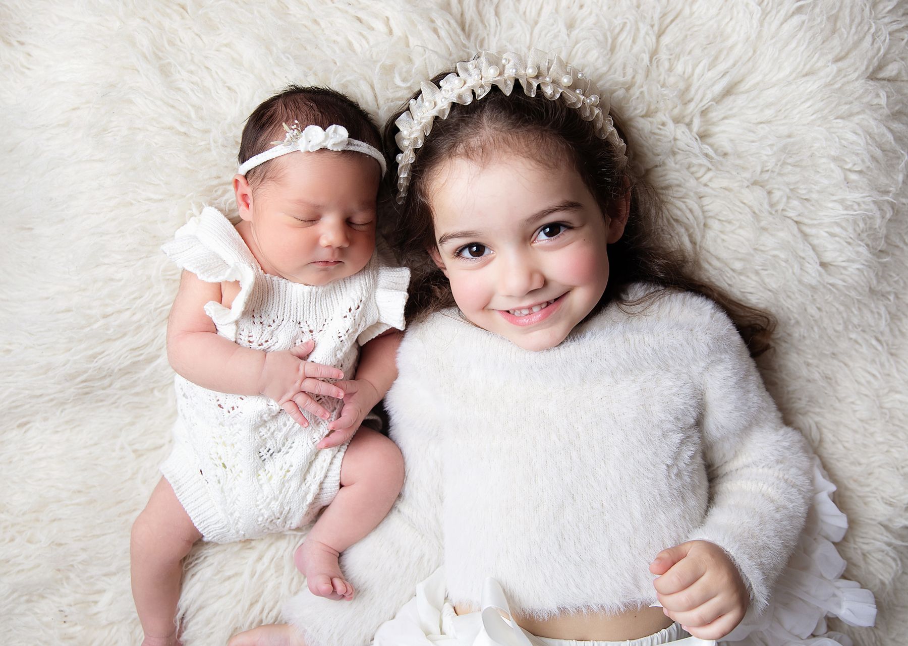 Two siblings lying on white fur, wearing white outfits and delicate headbands in a sweet studio portrait. During a newborn session In London
