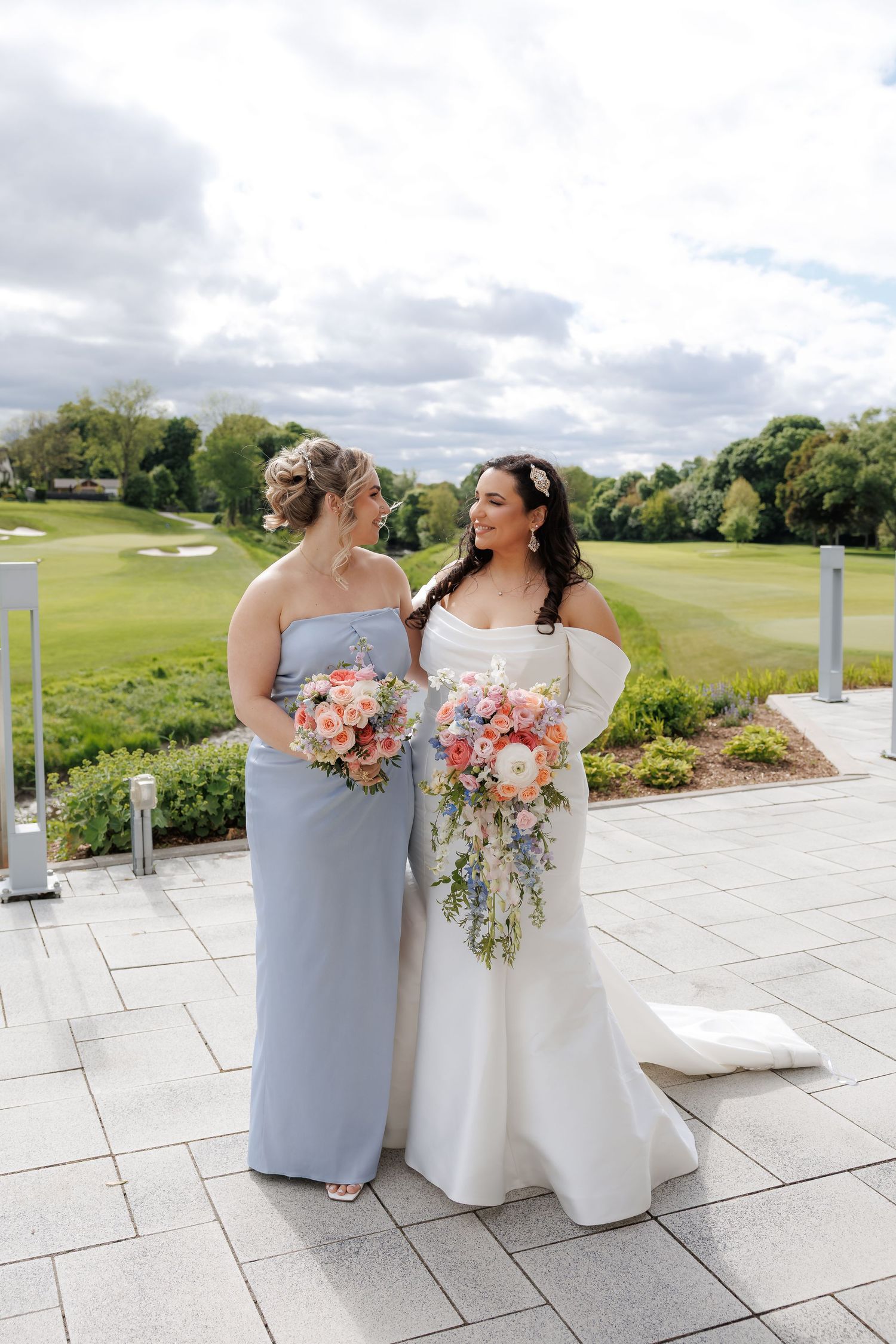 A bride and bridesmaid holding matching coral bouquets on a stone patio.