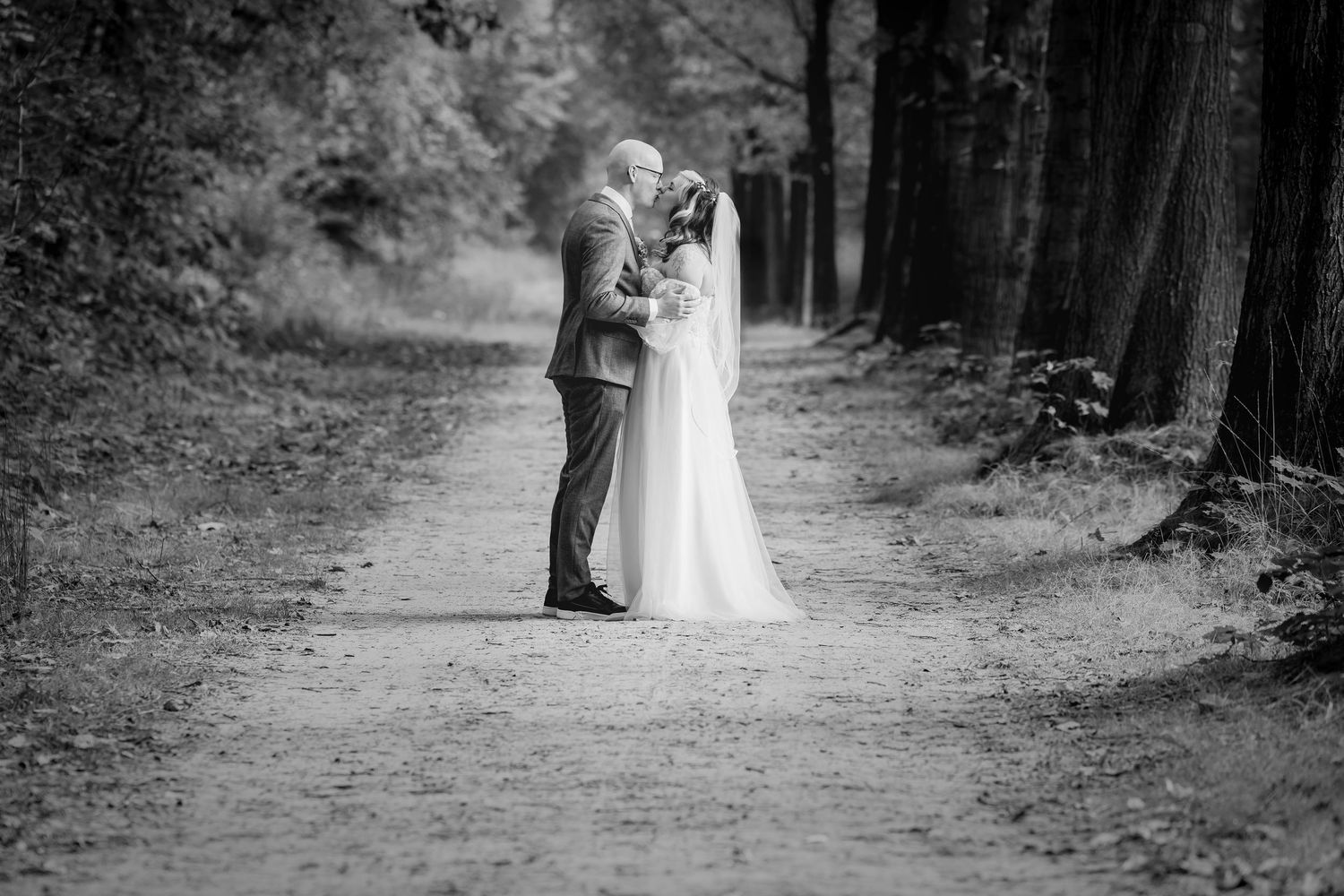 A romantic black and white wedding photo of a couple embracing on a forest path surrounded by tall trees.