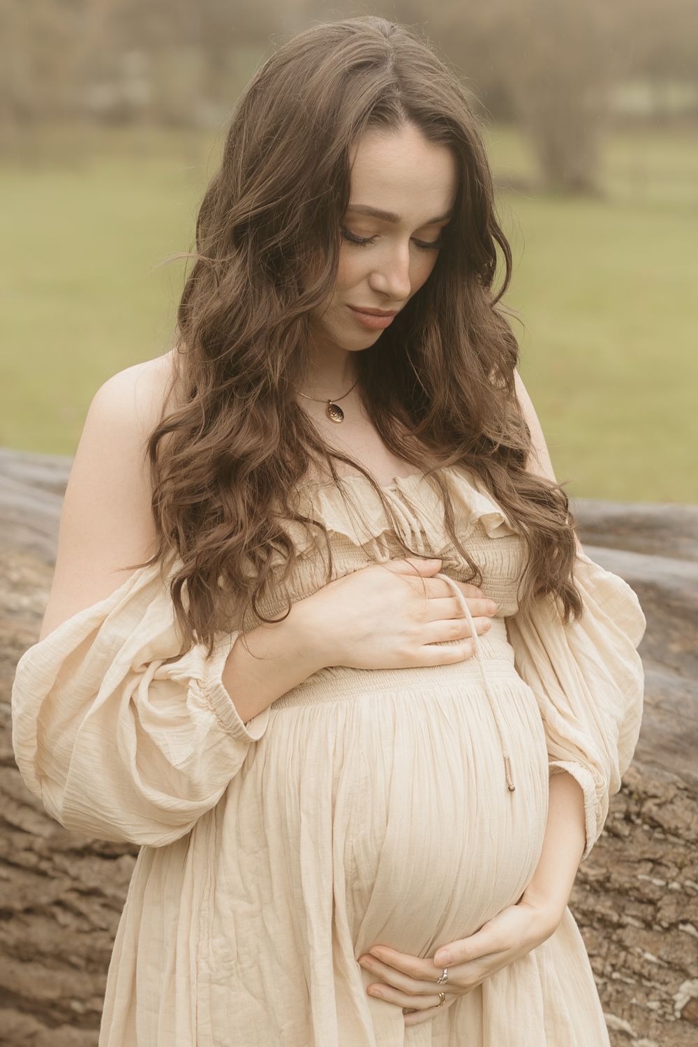 A pregnant woman in a flowy cream dress cradles her baby bump while standing outdoors in a dreamy natural setting.