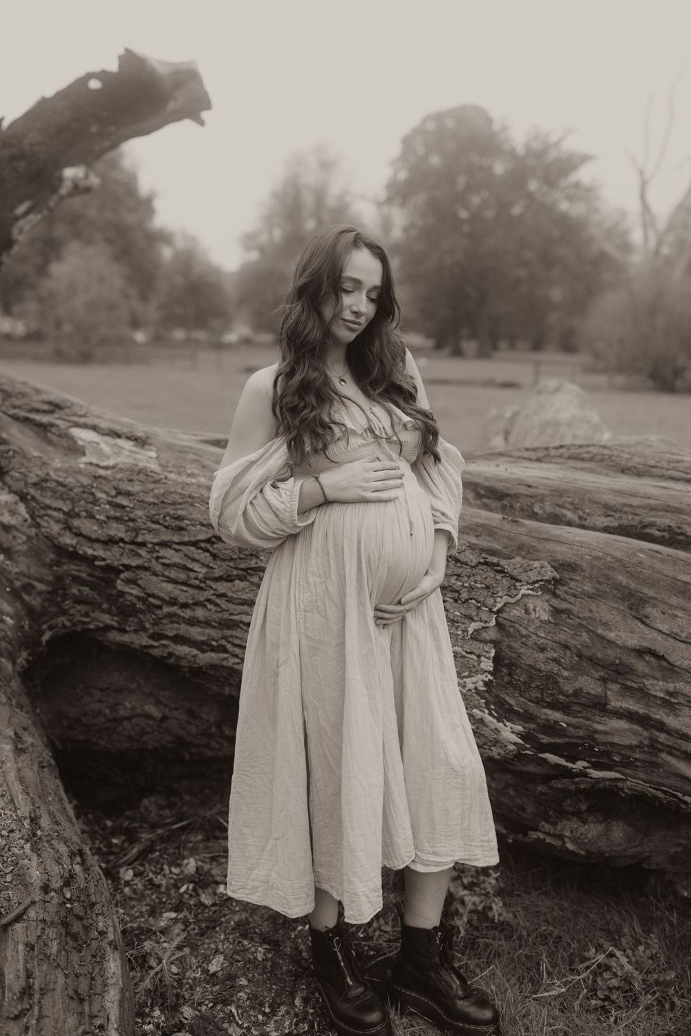 Black and white portrait of an expectant mother in a flowing white dress standing on a fallen log in a misty outdoor setting.